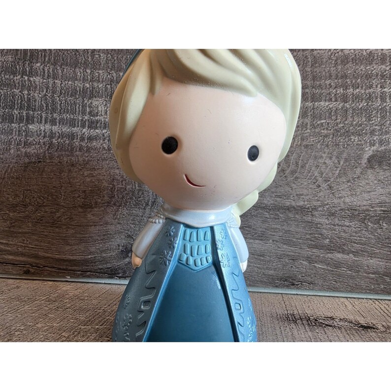 May include: A figurine of a cartoon character with blonde hair and a blue dress. The figure has a smiling face and is standing upright. The dress has a detailed design.