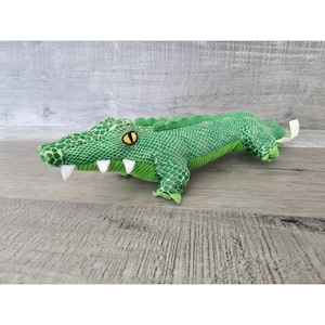 Peut inclure: Jouet en peluche d'alligator vert avec un design texturé et écailleux. L'alligator a un œil jaune et des dents blanches.