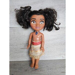 May include: A brown-skinned doll with long, curly black hair styled in pigtails. The doll is wearing a brown and gold skirt with a red waistband and a blue necklace with a pendant.