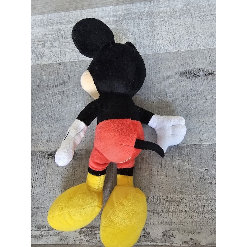 Puede incluir: Un juguete de peluche de Mickey Mouse con orejas negras, cuerpo negro, pantalones cortos rojos y zapatos amarillos.
