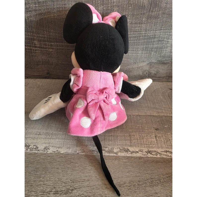 May include: A plush Minnie Mouse doll, seen from the back, with a pink dress adorned with white polka dots and a large bow. The doll has black ears, a pink and white polka dot bow, and white gloves. A black tail extends downwards.