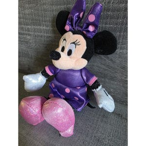 Op de afbeelding: Een zittende Minnie Mouse knuffel. Ze draagt een paarse jurk met roze stippen, een bijpassende strik en roze glitter schoenen. Haar zwarte oren en gezicht zijn zichtbaar, met witte handschoenen en een lieve uitdrukking.