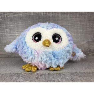 Può includere: Un giocattolo a forma di gufo di peluche con un corpo rotondo e grandi occhi espressivi. Il gufo presenta un mix di morbida pelliccia blu, viola e bianca, con accenti glitterati dorati sul becco e sulle zampe. Il giocattolo è morbido e coccoloso.