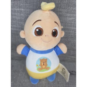 May include: A baby-shaped plush doll with a smiling face, brown eyes, and a small tuft of yellow hair. The doll is dressed in a white shirt with a bear graphic, blue sleeves, and yellow trim. A tag is attached to the doll.