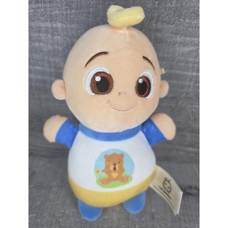 May include: A baby-shaped plush doll with a smiling face, brown eyes, and a small tuft of yellow hair. The doll is dressed in a white shirt with a bear graphic, blue sleeves, and yellow trim. A tag is attached to the doll.