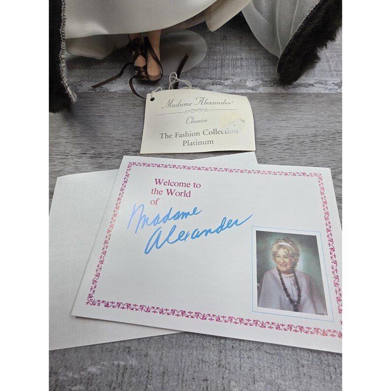 May include: A doll with a tag that reads "Madame Alexander Classics The Fashion Collection Platinum." A welcome card with the text "Welcome to the World of Madame Alexander" and a photo of a woman is also visible.