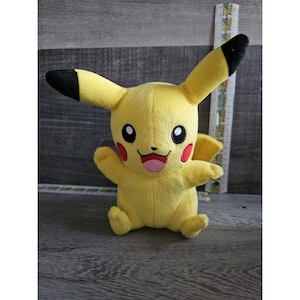 May include: A yellow plush toy of Pikachu, a popular character from the Pokemon franchise. The toy has black ears, a red nose, and black eyes. It is sitting on a wooden surface.