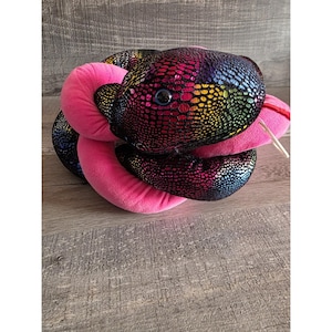 Può includere: Un giocattolo a forma di serpente di peluche con un motivo a scaglie color arcobaleno sulla testa e sul corpo. Il serpente ha un corpo rosa e una lingua rossa. Il giocattolo è arrotolato e sembra morbido e coccoloso.