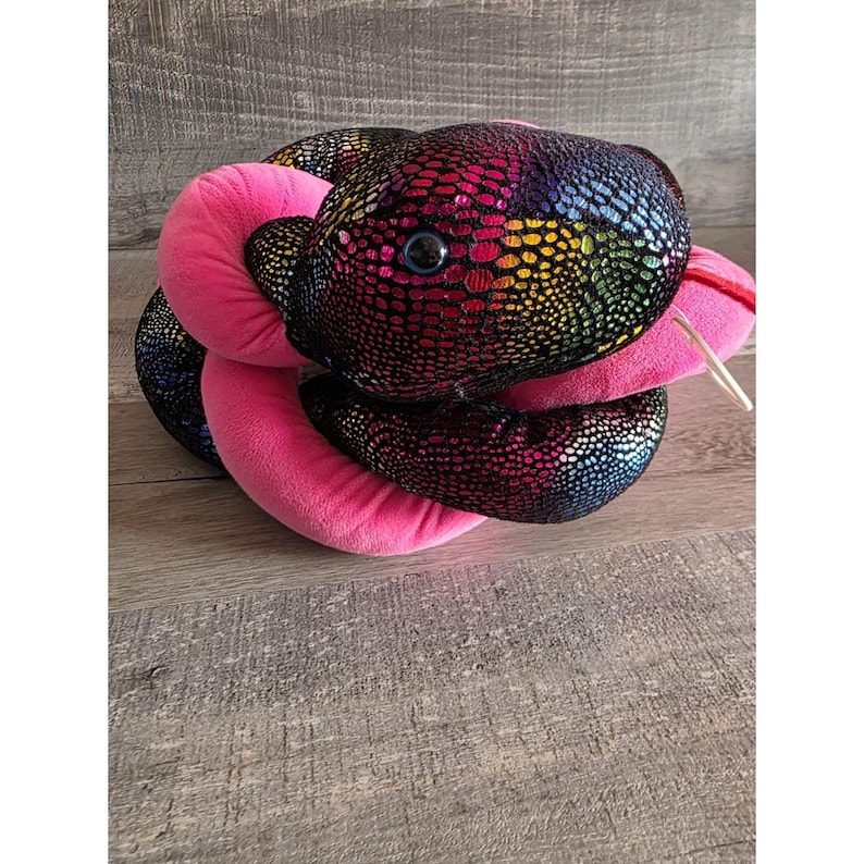 Op de afbeelding: Een pluche slangenspeelgoed met een regenboogkleurig, schubachtig patroon op zijn kop en lichaam. De slang heeft een roze lichaam en een rode tong. Het speelgoed is opgerold en ziet er zacht en knuffelig uit.
