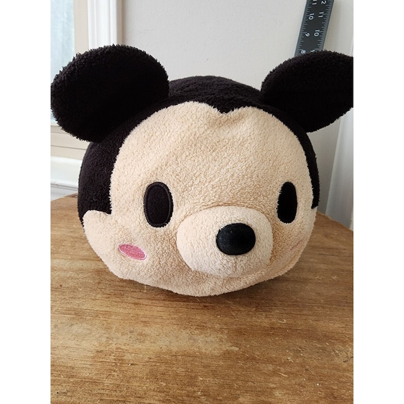 May include: A plush Mickey Mouse toy with black ears, a brown nose, and black eyes. The toy has a beige face with pink cheeks.