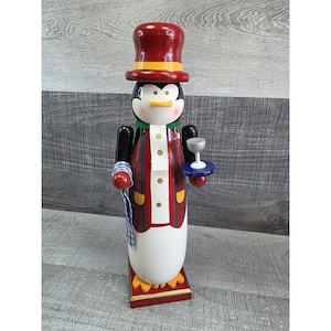 May include: A wooden penguin figurine wearing a red top hat with a yellow band, a red and green striped jacket, and a white shirt. The penguin holds a small silver goblet on a blue tray and a rolled-up napkin. The figurine stands on a red base.
