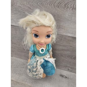 May include: A blonde haired doll wearing a blue and white dress with a snowflake design. The doll has a white necklace with a snowflake pendant.