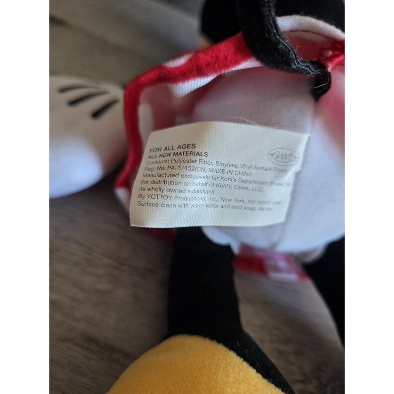 May include: A plush Minnie Mouse doll with a red and white polka dot bow. The doll has black ears, a white face, and a tag that reads "FOR ALL AGES ALL NEW MATERIALS". The doll's features include a black nose and a yellow and black outfit.