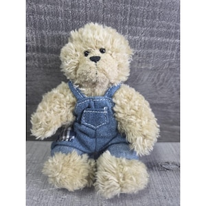 Op de afbeelding: Een teddybeer met lichtbruine vacht en een zwarte neus. De beer draagt een blauwe spijkerbroek met witte stiksels en een zak. De tuinbroek heeft een ruitpatroon op de rug.