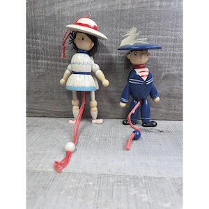 May include: Two wooden pull-string dolls. One doll is dressed in a white dress with blue trim and a white hat with a red band. The other doll is dressed in a blue sailor suit with a red and white striped neckerchief and a blue hat with a white feather.