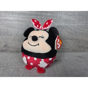 May include: A round plush toy resembling a cartoon mouse with black ears, a cream-coloured face, and a red dress with white polka dots. It features a red and white polka-dot bow on top, white hands, red feet, and a heart-shaped Ty brand tag.