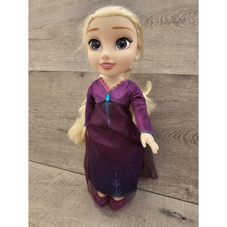 May include: A purple and blue dress with a sparkly design and a blue gem on the bodice. The doll has blonde hair and blue eyes.