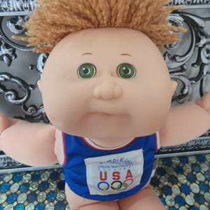 May include: A doll with light skin, green eyes, and short, textured light brown yarn hair. The doll wears a blue sleeveless top with white and red trim, featuring a white patch that reads Cabbage Patch Kids Official Team Mascot USA.