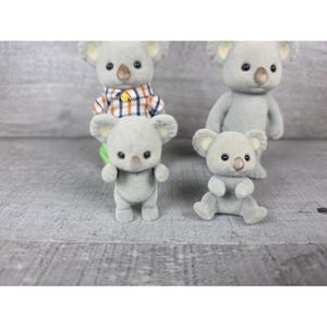 May include: Four gray koala figurines with black eyes and tan noses are arranged on a gray surface. One koala wears a plaid shirt. One koala is sitting, and the others are standing. The koalas have large, round ears.