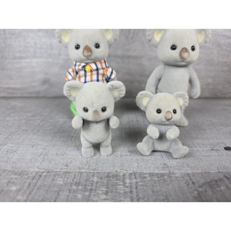 May include: Four gray koala figurines with black eyes and tan noses are arranged on a gray surface. One koala wears a plaid shirt. One koala is sitting, and the others are standing. The koalas have large, round ears.