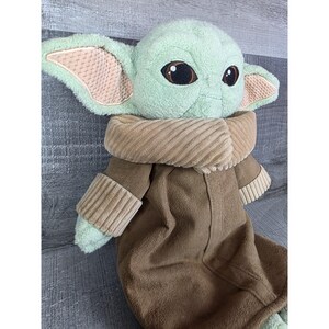 May include: A plush doll of a character with large ears, big eyes, and a brown robe. The doll has a light green face and ears with a tan interior. The robe is brown with a tan collar and sleeves. The doll is lying on a gray surface.