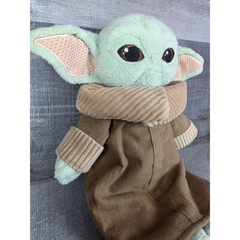 May include: A plush doll of a character with large ears, big eyes, and a brown robe. The doll has a light green face and ears with a tan interior. The robe is brown with a tan collar and sleeves. The doll is lying on a gray surface.