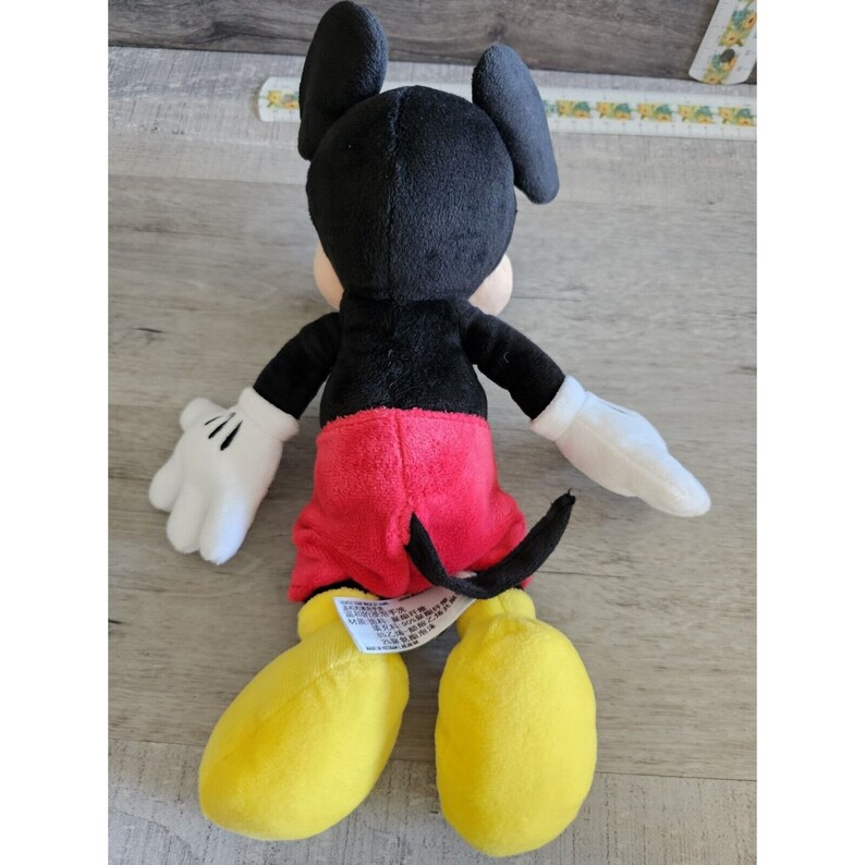 Puede incluir: Un juguete de peluche de Mickey Mouse con pelaje negro, pantalones cortos rojos, guantes blancos y zapatos amarillos. El juguete est&aacute; sentado sobre una superficie de madera.