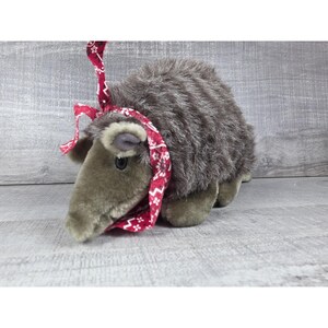 May include: A plush stuffed animal resembling an armadillo with shaggy dark gray fur and a smooth light gray snout. The creature is wearing a red fabric bonnet tied under its chin, featuring a white floral and geometric pattern throughout the fabric.