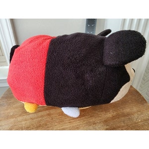 May include: A plush toy shaped like Mickey Mouse. The toy is red, black, and tan. The toy is lying on a wooden surface.