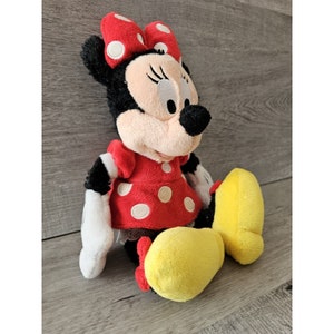 K&ouml;nnte beinhalten: Ein Pl&uuml;schtier von Minnie Mouse, die ein rotes Kleid mit wei&szlig;en Punkten und eine rote Schleife mit wei&szlig;en Punkten tr&auml;gt. Sie hat schwarze Augen, Wimpern und eine schwarze Nase. Ihre Schuhe sind gelb.