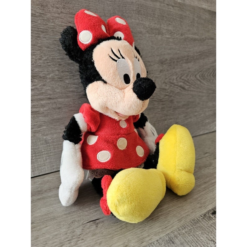K&ouml;nnte beinhalten: Ein Pl&uuml;schtier von Minnie Mouse, die ein rotes Kleid mit wei&szlig;en Punkten und eine rote Schleife mit wei&szlig;en Punkten tr&auml;gt. Sie hat schwarze Augen, Wimpern und eine schwarze Nase. Ihre Schuhe sind gelb.