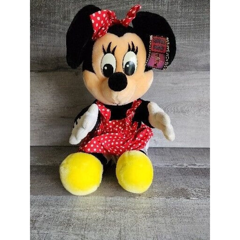 Pu&ograve; includere: Un giocattolo di peluche di Minnie Mouse, la fidanzata di Topolino. Indossa un vestito rosso con pois bianchi e un fiocco nero. Ha un viso giallo con occhi neri e un naso nero. &Egrave; seduta su una superficie di legno.
