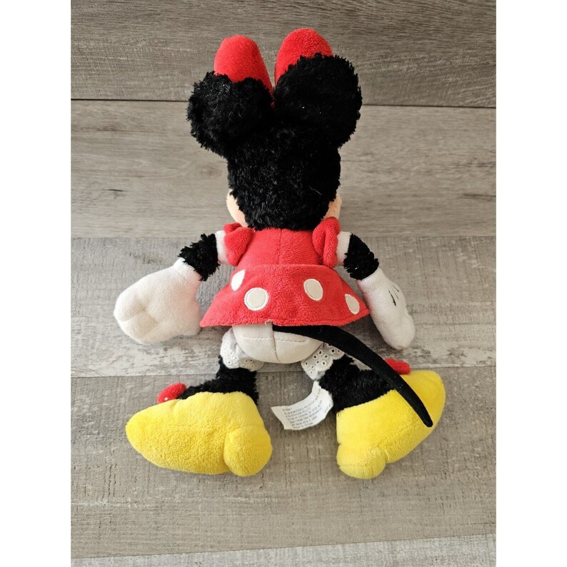 K&ouml;nnte beinhalten: Ein Pl&uuml;schtier von Minnie Mouse, die ein rotes Kleid mit wei&szlig;en Punkten und gelbe Schuhe tr&auml;gt. Sie hat schwarzes Fell und einen schwarzen Schwanz.