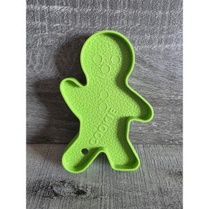 May include: A bright green gingerbread man cookie cutter. The cutter has a textured surface and the word "COOKIE" is printed on the front. The cutter has a hole at the bottom.