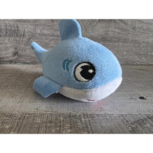 May include: A plush toy shark in light blue with a white belly and a small red mouth. The shark has large, expressive eyes with black pupils and embroidered details. The toy is designed with fins and a tail.