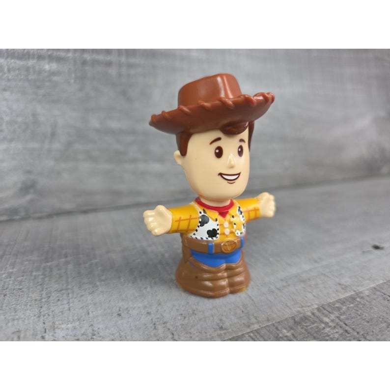 May include: A plastic toy figure of Woody from Toy Story. The figure has a brown cowboy hat, a yellow shirt with a cowhide pattern, and brown pants. The figure's arms are outstretched.