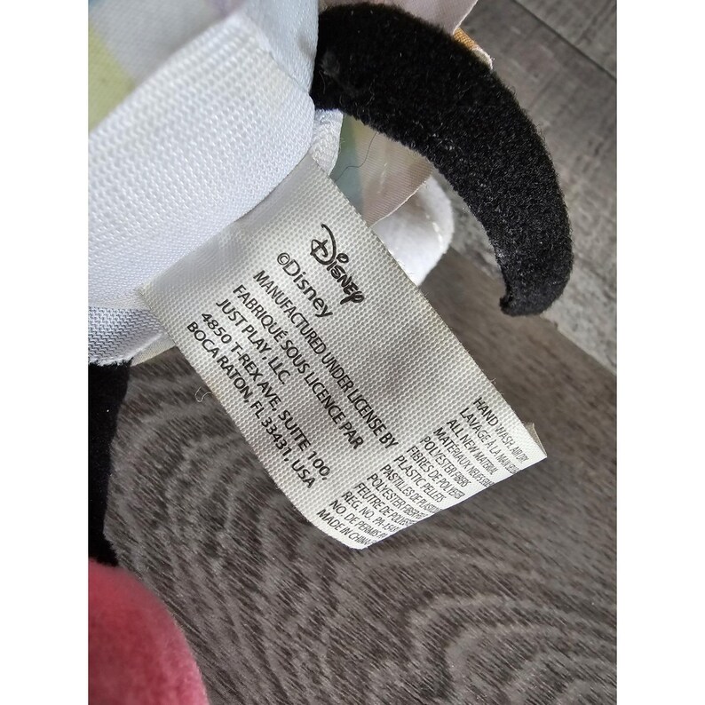 K&ouml;nnte beinhalten: Ein wei&szlig;es Etikett mit schwarzem Text, der "&copy;Disney", "Manufactured under license by Just Play, LLC", "Fabriqu&eacute; sous licence par Just Play, LLC", "4850 T-Rex Ave, Suite 100, Boca Raton, FL 33431, USA", "Hand Wash/Lavage &agrave; la Main", "All New Materials/Mat&eacute;riaux Neuf", "Polyester Fibers/Fibres de Polyester", "Plastic Pellets/Pastilles de Plastique", "Polyester Fiber/Fibre de Polyester", "Felt/Feutre de Poulie", "Reg. No. PA-1", "No. de Permis", "Made in China/Fabriqu&eacute; en Chine" lautet.