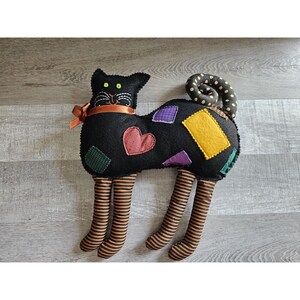 May include: Black felt cat with a patchwork design of colorful felt squares and hearts. The cat has a long, curled tail and striped legs.