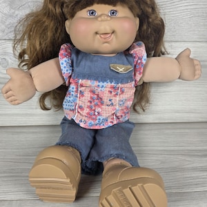 Könnte beinhalten: Eine Puppe mit braunen Zöpfen, blauen Augen und einem runden Gesicht. Die Puppe trägt ein blaues Oberteil im Jeans-Look mit einem rosa-blauen Blumenmuster auf der Brust, einer kleinen Tasche und einer passenden blauen Jeanshose. Beige Schuhe mit CPK.
