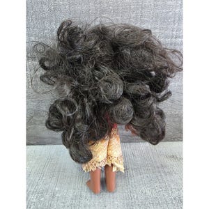 Op de afbeelding: Een pop met donkerbruin, krullend haar en een beige en gouden rok. De huid van de pop is bruin en de pop staat op een grijs oppervlak.
