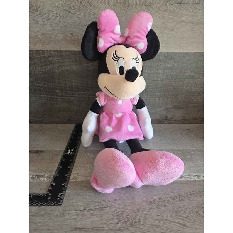 May include: A plush Minnie Mouse doll, featuring a pink dress with white polka dots and a matching bow. The doll has black ears, a white face, and large pink feet. The doll is approximately 18 inches tall.