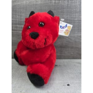 Pode incluir: Um brinquedo de diabo de pelúcia vermelho com chifres, nariz e pés pretos. O bicho de pelúcia tem olhos grandes e redondos e um pequeno sorriso. Uma etiqueta branca está presa ao brinquedo.