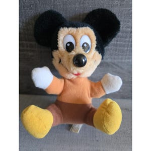 Puede incluir: Un peluche de Mickey Mouse con orejas negras, cara bronceada y nariz negra. El juguete tiene un torso naranja, pantalones marrones, guantes blancos y pies amarillos. El juguete está sentado con las piernas extendidas.