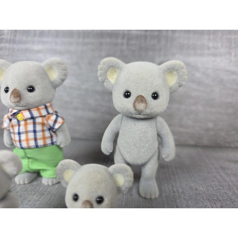 May include: Several gray koala figurines with black eyes and brown noses are displayed. One koala wears a plaid shirt and green pants. The koalas have large ears with yellow inner ears. The figurines are on a gray surface.