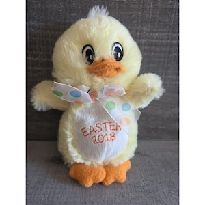 May include: A plush yellow chick with orange feet and beak, and large black eyes. It has a white belly with the embroidered text "EASTER 2018". A sheer bow with colorful polka dots is tied around its neck. A cute Easter decoration.