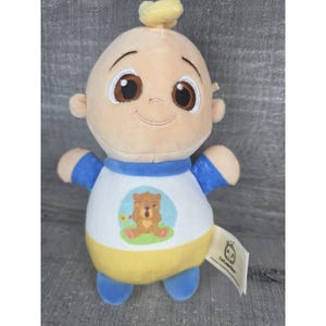 May include: A soft toy baby doll with a smiling expression, brown eyes, and a small yellow hair tuft. The doll is wearing a white shirt with a bear design, blue sleeves, and a yellow lower section. A tag with a logo is attached.
