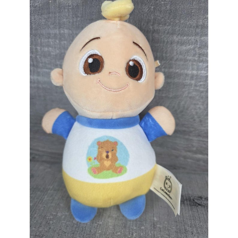 May include: A soft toy baby doll with a smiling expression, brown eyes, and a small yellow hair tuft. The doll is wearing a white shirt with a bear design, blue sleeves, and a yellow lower section. A tag with a logo is attached.