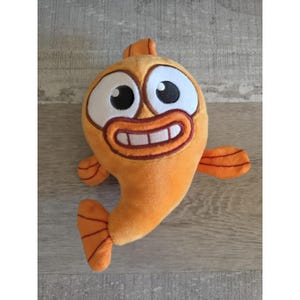 May include: Orange plush fish toy with large white-rimmed eyes, a wide grin, and small fins. The cartoon-style fish has a cheerful expression and is made of soft, fuzzy material. A fun, whimsical toy for children.