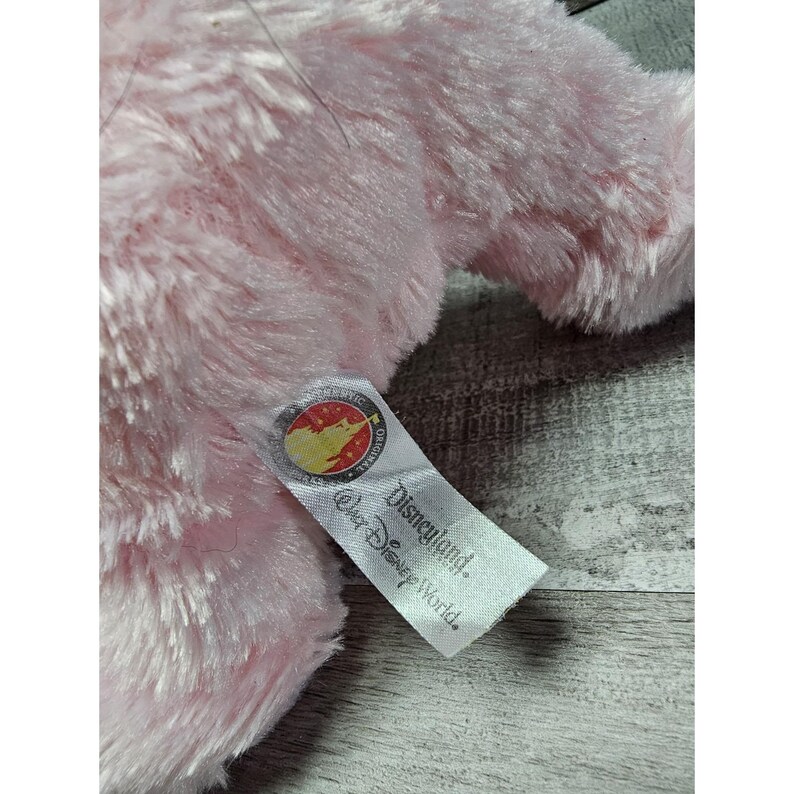 May include: A close-up of a pink plush toy with a tag that reads "Disneyland. Walt Disney World." The tag has a circular emblem with a red and yellow design. The plush toy has a soft, fluffy texture.