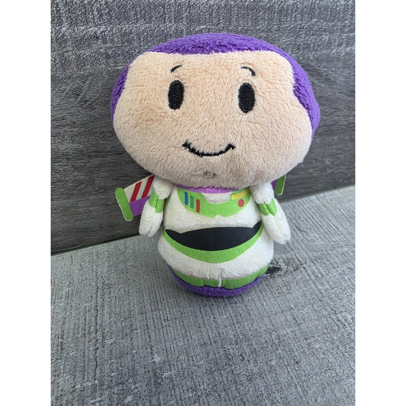May include: A plush toy of the character Buzz Lightyear from the Toy Story franchise. The toy has a rounded head with purple hair, a smiling face, and a white and green spacesuit. The toy is approximately 6 inches tall.